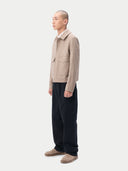 A man wearing a light beige cashmere utility jacket with a collar, front button closure, and two large flap chest pockets, layered over a white collared shirt and dark trousers, against a plain light gray studio background. #color_taupe
