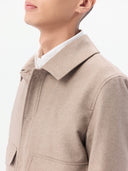 A man wearing a light beige cashmere utility jacket with a collar, front button closure, and two large flap chest pockets, layered over a white collared shirt and dark trousers, against a plain light gray studio background. #color_taupe