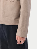 A man wearing a light beige cashmere utility jacket with a collar, front button closure, and two large flap chest pockets, layered over a white collared shirt and dark trousers, against a plain light gray studio background. #color_taupe