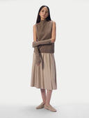A model stands against a light gray studio background wearing a sleeveless taupe-brown mock-neck knit sweater vest, a beige pleated midi skirt, matching beige knit arm warmers, and beige loafers.