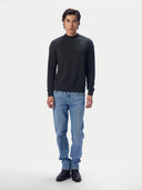A man modeling a dark charcoal gray mock neck knit sweater, paired with light blue jeans and black leather boots, against a plain white studio background.
