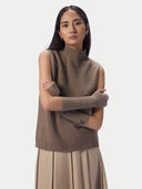 A model wearing a taupe brown sleeveless mock-neck knit sweater vest with matching ribbed knit arm warmers, paired with a pleated beige skirt, against a light gray studio background.