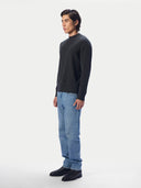 A man wearing a dark charcoal gray knit mock-neck sweater, light blue jeans, and black leather shoes, standing in profile against a light gray studio background.