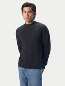 A man wearing a dark charcoal gray knit crewneck sweater and blue jeans, standing against a light gray studio background.