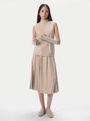 A model wearing a beige knit turtleneck sweater vest with matching ribbed arm warmers and a pleated midi skirt, paired with beige slip-on shoes, against a light gray studio background.