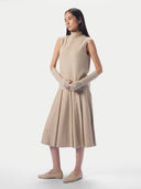 A model wearing a beige knit sweater vest, matching ribbed knit arm warmers, a pleated beige midi skirt, and beige loafers, standing against a light gray studio background.