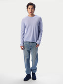 A man wearing a light blue long-sleeve crewneck sweater, light-wash blue jeans and dark navy suede shoes, standing against a light gray studio background.