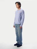 A man shown in side profile wearing a light blue long-sleeve crewneck sweater, light-wash blue jeans and dark navy suede shoes, against a light gray studio background.