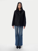 A model stands against a light gray studio background wearing a black button-front jacket, blue flare jeans, and black pointed shoes.