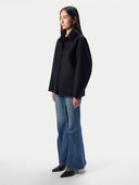 A woman stands in profile wearing a black button-front overshirt with a collar, blue wide-leg jeans, and black pointed shoes against a clean light gray studio background.