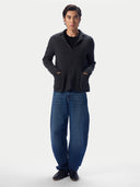 A man modeling a dark charcoal gray knit cardigan with front pockets over a black shirt and loose blue jeans, standing against a light gray studio background.
