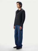 A man stands in profile wearing a dark charcoal gray knit cardigan over a black shirt, paired with blue jeans and black shoes, against a plain light gray studio background.