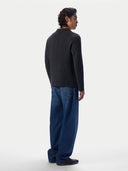 A man shown from the back wearing a dark charcoal gray knit sweater and loose blue jeans, standing against a plain light gray studio background.