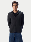 A man wearing a dark charcoal gray knit cardigan over a black t-shirt and blue jeans, standing against a light gray studio background.