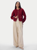 A woman stands against a light gray studio background wearing a burgundy cropped cardigan with decorative trim and cream wide-leg pants.