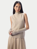 A model wearing a beige sleeveless mock-neck knit sweater vest with matching ribbed knit arm warmers and a pleated beige skirt, set against a solid light gray background.