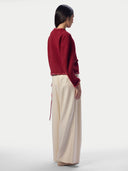 A model shown from the back wearing a deep red cropped knit sweater with red beaded trim and cream wide-leg trousers, set against a plain light gray studio background.