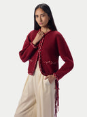 A woman modeling a cropped, long-sleeve burgundy knit cardigan with decorative braided trim and fringe details, paired with cream wide-leg pants, against a light gray studio background.
