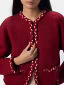 A close-up of a model wearing a deep red knit cardigan with braided trim details in red and white along the collar, front opening, cuffs, and pockets.