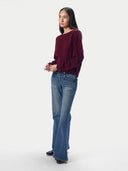 A woman modeling a long-sleeve boat-neck knit top in deep burgundy, paired with blue flare jeans and black shoes, standing against a light gray studio background.