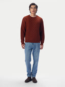 A man wearing a rust-brown cable-knit sweater, blue jeans, and dark brown shoes, standing against a plain light gray studio background.