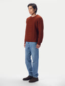 A man wearing a rust-brown cable-knit crewneck sweater, blue jeans, and dark brown suede boots, standing against a plain light gray studio background.