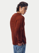 A man wearing a rust-brown cable-knit sweater and blue jeans, shown from a side-back angle against a light gray studio background.