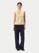 A model stands against a light gray studio background wearing a pale yellow sleeveless mock-neck knit sweater vest and dark charcoal wide-leg trousers.