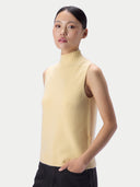 A woman wearing a pale yellow sleeveless mock-neck knit sweater, paired with black pants, photographed against a light gray studio background.