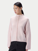 A woman modeling a pale pink knit mock-neck sweater with a decorative braided front detail, paired with black pants, against a light gray studio background.