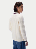 A man wearing a cream cable-knit sweater with striped collar and cuffs, paired with blue jeans, shown from a side-back angle against a light gray studio background.
