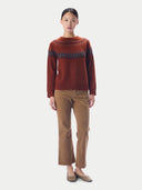 A model stands against a light gray studio background wearing a rust-brown knit sweater with a dark blue Nordic pattern across the chest, paired with brown corduroy trousers and brown shoes.