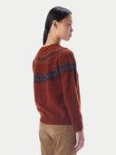 A woman shown from the back wearing a rust-orange knit sweater with a dark blue and gray geometric pattern across the shoulders and upper back, paired with tan pants, against a light gray studio background.
