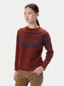 A woman modeling a rust-brown knit sweater with a dark blue geometric pattern across the chest, paired with brown pants, against a light gray studio background.
