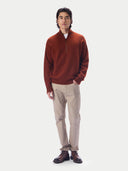 A man modeling a rust-orange quarter-zip knit sweater, paired with beige trousers and brown leather shoes, standing against a solid light gray background.