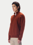 A man modeling a rust-orange quarter-zip knit sweater, paired with beige trousers and brown leather shoes, standing against a solid light gray background.