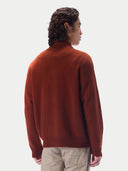 A man shown from the back wearing a rust-orange knit sweater and light beige trousers, photographed against a clean off-white studio background.