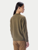 A woman shown from the back wearing a textured olive green knit sweater with cable detailing on the shoulders and sides, paired with brown pants, against a light gray studio background.