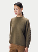 A woman wearing a muted olive-brown knit sweater with a textured cable pattern on one sleeve, paired with brown pants, against a light gray studio background.