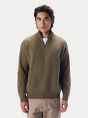A man wearing a muted olive-brown quarter-zip knit sweater over a white undershirt and beige trousers, standing against a plain light gray studio background.#color_muted-green