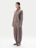 A model stands in profile wearing a matching taupe knit two-piece outfit consisting of a mock-neck long-sleeve top with a braided front detail and wide-leg pants, paired with flat brown shoes, against a plain light gray studio background.