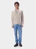 A man modeling a light beige zip-up knit cardigan over a white t-shirt, paired with blue jeans and dark brown suede shoes, against a solid light gray background.#color_natural-beige