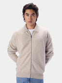 A man wearing a light beige zip-up knit cardigan over a white t-shirt and blue jeans, photographed against a solid light gray background.#color_natural-beige
