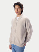 A man wearing a light beige zip-up knit sweater over a white t-shirt and blue jeans, photographed against a solid light gray background.#color_natural-beige
