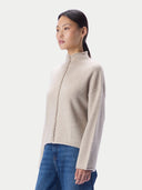 A woman wearing a light beige knit mock-neck sweater with a braided front detail and blue jeans, photographed against a clean light gray studio background.