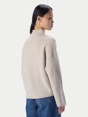 A woman shown from the back wearing a light beige mock-neck knit sweater and blue jeans against a plain light gray studio background.