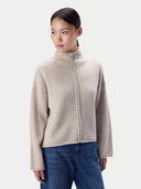 A woman wearing a light beige knit mock-neck sweater with a braided front detail, paired with blue jeans, against a solid light gray background.