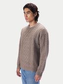 A man wearing a taupe cable-knit crewneck sweater and light blue jeans, photographed from the waist up against a light gray studio background.