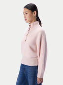 A woman in profile wearing a light pink ribbed mock-neck sweater with brown buttons and blue jeans, photographed against a clean light gray studio background.