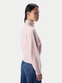 A woman shown in side profile wearing a light pink ribbed turtleneck sweater and blue jeans against a plain light gray studio background.